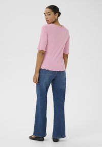 Roze ribfluwelen top met gegolfde randen, gecombineerd met hooggewaist blauwe jeans. Model staat met haar rug naar de camera en toont de achterzijde van de kleding.