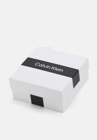 Scatola regalo quadrata bianca con una fascia nera, con il testo "Calvin Klein". Superficie liscia, design pulito, dimensioni compatte.