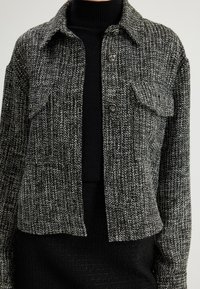 Zwart-witte textuurjack met een knoopsluiting aan de voorkant, een borstzak en een puntige kraag. Materiaal heeft een geweven patroon.