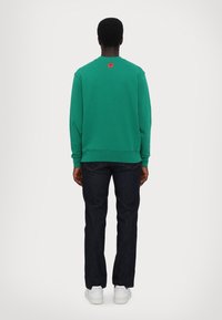 Grøn bomulds sweatshirts med rund halsudskæring, ribbede manchetter og kant. Har et lille rødt logo på bagsiden. Bæres med mørke jeans og hvide sneakers.