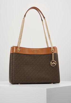 Brązowa i beżowa torebka Michael Kors z złotymi łańcuszkowymi paskami i zawieszką z logo MK, spoczywająca na białym piedestale na jednolitym tle.