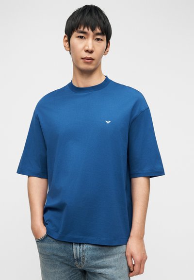 Νέος άνδρας που φοράει μπλε κοντομάνικο πουκάμισο με μικρό λευκό λογότυπο στο στήθος, στέκεται με το ένα χέρι στην τσέπη του τζιν.