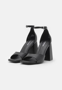 Sandales à talons hauts noires avec bout ouvert, bride de cheville et talons bloc. Fabriquées en cuir lisse, elles présentent un design élégant et un minimum de détails.