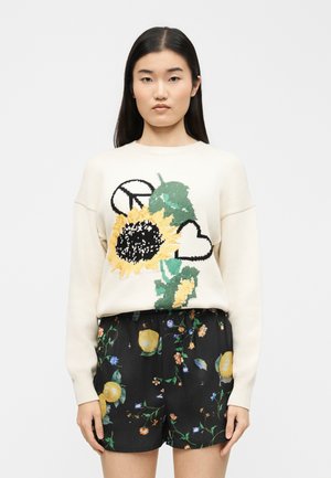 Naine kannab kreemikat kampsunit päevalille-, südame- ja rahumärgi mustritega, kusjuures tal on seljas mustad lillemustriga püksid.