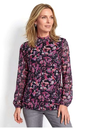 Frau trägt eine langärmelige Blumenbluse mit lila und pinken Mustern und graue Hose, schaut seitlich vor weißem Hintergrund.