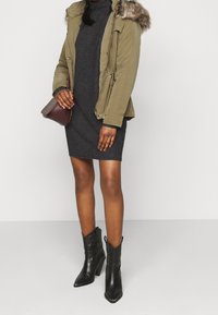 Parka verde oliva con bordo in pelliccia sintetica, chiusura con zip e tasche laterali, abbinata a un vestito di maglia grigio scuro e stivali alla caviglia neri con punta a stiletto.
