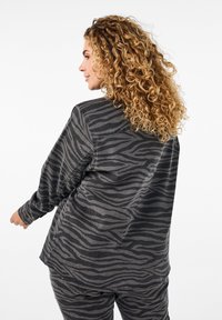 Kvinde med langt krøllet hår iført matchende mørk zebra-stribet jakke og bukser, drejet let til siden mod en hvid baggrund.