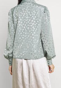 Lysgrøn bluse med en glat, tekstureret overflade med sølvfarvet leopardprint, lange puffede ærmer og knaplukning ved håndleddene.