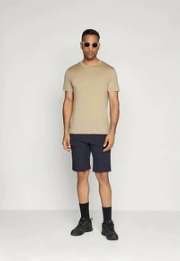 Bež majica s kratkimi rokavi, temni kratki hlače, črne nogavice in črne športne copate. Model nosi svetle sončne očala in stoji pred enostavno ozadje.