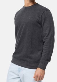 Ciemnoszary sweatshirt z okrągłym dekoltem, długimi rękawami i prążkowanymi mankietami. Subtelny detal logo na piersi. Miękka tekstura, casualowy design.
