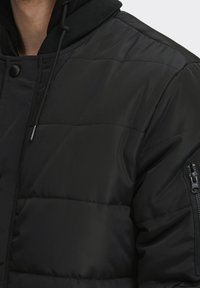 Czarna kurtka puchowa z pikowanym wzorem, wyposażona w zamykaną na zamek kieszeń na lewym rękawie oraz kaptur z czarnymi sznureczkami.