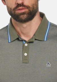 Olivgrünes Poloshirt mit strukturiertem Muster, drei Knöpfen und einem blau gestreiften Kragen. Mit kleinem Pinguin-Logo auf der Brust.