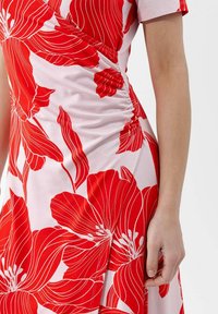 Rotes Blumenkleid mit großen roten Blumen und weißen Konturen auf einem hellrosa Hintergrund, mit gerafftem Material an der Taille und kurzen Ärmeln.