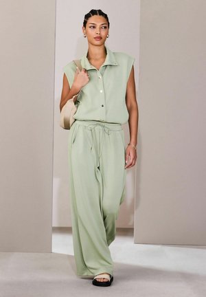 Femeie cu părul împletit, purtând o cămașă fără mâneci, cu nasturi, de culoare verde deschis, și pantaloni largi, purtând o geantă bej pe umăr și sandale.