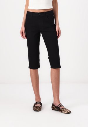 Person trägt schwarze knielange Hose, weißes Cropped-Top und flache Schuhe mit Leopardenmuster, steht vor weißem Hintergrund.
