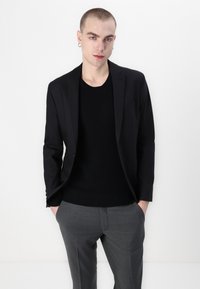 Fekete blazer fekete pulóver fölött, szürke nadrággal párosítva. Simaság jellemzi az anyagot, igényes szabás, minimális részletezés és éles sziluett.