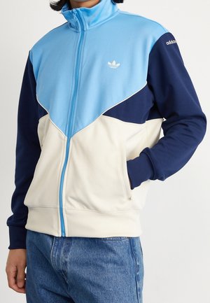 Osoba ubrana w niebieską, granatową i kremową kurtkę na zamek Adidas z logo na piersi i rękawie, w połączeniu z niebieskimi jeansami.