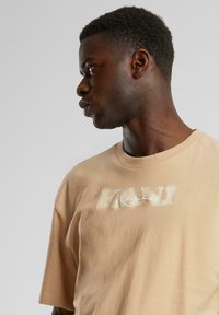 Beige oversized t-shirt featuring a faded white logo on the chest, short sleeves, and a crew neckline. Soft cotton material.