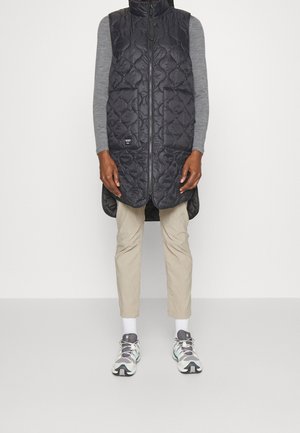 Personne portant une longue veste matelassée noire sur un chemisier gris à manches longues, un pantalon beige court, des chaussettes blanches et des chaussures de sport grises, debout à l'intérieur.