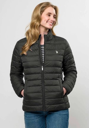 Woman smiles looking sideways wearing a black quilted jacket with hands in pockets and blue jeans against a plain light background.
