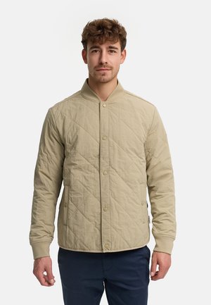 Ung mand iført en beige quiltet jakke med trykknapper og marineblå bukser, stående foran en ensfarvet hvid baggrund.