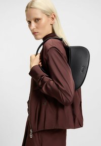 Mochila de cuero negra con una forma aerodinámica, que presenta un diseño curvado y una marca minimalista, llevada al hombro. Textura suave.