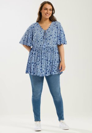 Uśmiechnięta kobieta w niebieskiej tunice w kwiaty z falbaniastymi rękawami, dopasowanych niebieskich dżinsach i białych tenisówkach stoi na tle jednolitego tła.