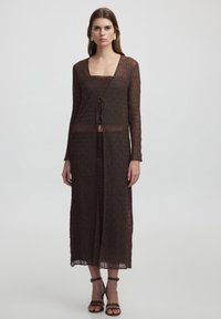 Braunes gestricktes Kleid mit langen Ärmeln, V-Ausschnitt und Kordelzugverschluss. Transparente Textur, knielange Länge, kombiniert mit braunen Riemchensandalen.