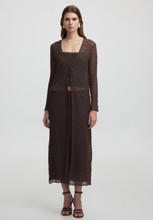 Brown knitted dress with long sleeves, V-neck, and drawstring closure. Sheer texture, mid-calf length, styled with brown strappy sandals.