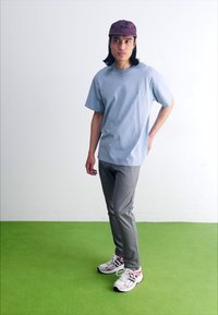 Világoskék, rövid ujjú póló, szürke slim-fit nadrág és fehér, piros részletekkel ellátott sportcipő. A modell zöld padlón áll, fehér fal előtt.