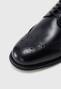 Μύτη μαύρου δερμάτινου υποδήματος brogue με διακοσμητικές διάτρητες λεπτομέρειες και κορδόνια σε λευκό φόντο.