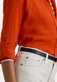 Bright orange linen shirt with buttoned cuffs, paired with white denim pants. Features a dark blue belt and contrasting white inner collar.