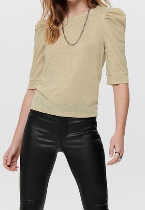 Kobieta ubrana w beżową błyszczącą bluzkę z bufkami i czarne błyszczące skórzane spodnie, uzupełniona łańcuszkowym naszyjnikiem i pierścionkami.