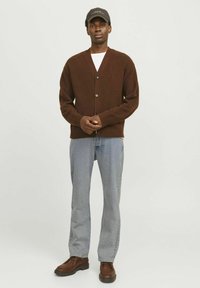 Cárdigan de punto marrón con escote en V y cinco botones, combinado con jeans azul claros y zapatos marrones de cordones. Acentuado con una gorra negra.