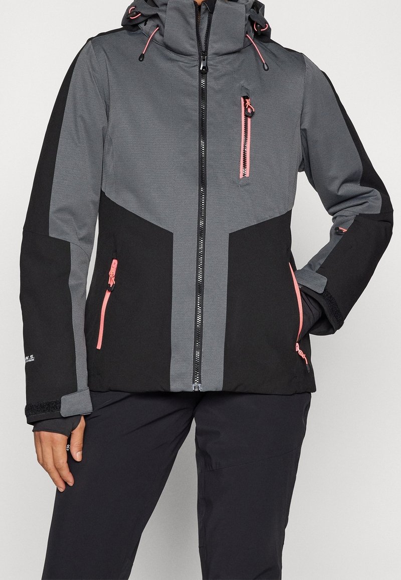 Femme portant une veste d'extérieur à capuche grise et noire avec des poches zippées roses et un pantalon noir, debout avec une main dans la poche.
