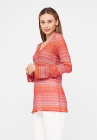 Langärmlige Tunika mit Zickzackmuster in Orange- und Rosatönen. V-Ausschnitt-Design mit leicht ausgestelltem Schnitt an den Hüften.