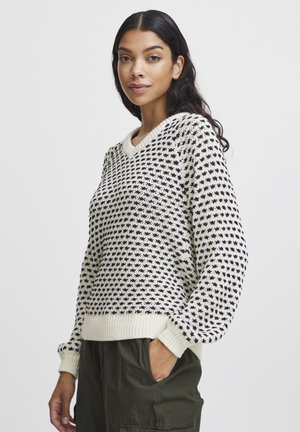 Schwarzer und weißer gestrickter Pullover mit V-Ausschnitt, ausgestattet mit einem strukturierten Sternmuster sowie gerippten Bündchen und Saum. Kombiniert mit olivgrünen Hosen.