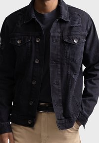 Hombre vistiendo una chaqueta de mezclilla negra desgastada sobre una camisa negra, pantalones beiges con un cinturón trenzado negro, con una mano en el bolsillo.