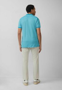 Türkisfarbenes Poloshirt mit kurzen Ärmeln und Kragen, kombiniert mit hellbeigen Hosen und weißen Sneakers. Glatte Stofftextur, lässige Passform.