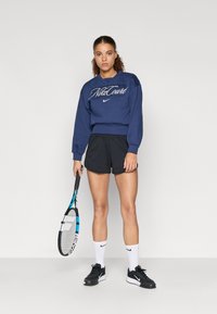 Ναυτικό sweatshirt με λογότυπο "NikeCourt", μαύρη βερμούδα και μαύρα αθλητικά παπούτσια. Κρατάει μια μπλε και λευκή ρακέτα τένις. Ορατές λευκές αθλητικές κάλτσες.