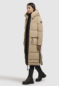 Beige puffjakke med høj krave, frontlynlås og sidelommer. Har sorte detaljer og et langt, lige design. Bæres med sorte støvler.