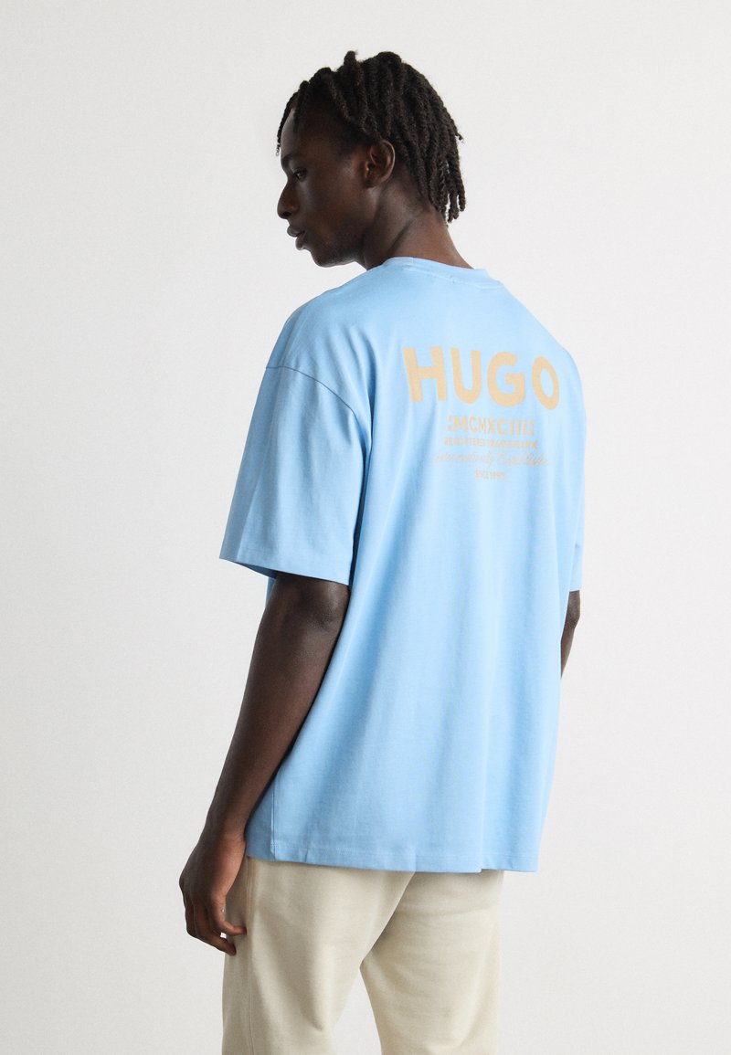 Svijetloplavi pamučni T-shirt s kratkim rukavima. Ima veliki kremasti natpis "HUGO" i dizajnerske elemente na leđima. Glatka tekstura, ležeran kroj.