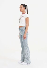 Lyseblå brede jeans med hvid blomsterbroderi, parret med en hvid cropped t-shirt med røde accenter og grafisk design. Hvite sneakers.