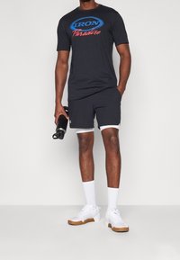 Czarna bawełniana koszulka z czerwonym i niebieskim logo, w połączeniu z ciemnymi szortami z białym obszyciem, białymi skarpetkami i kolorowymi butami sportowymi.