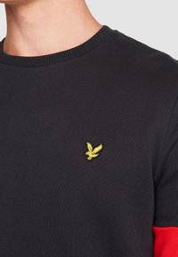 Sweatshirt nero a maglia con colletto tondo, caratterizzato da un logo ricamato giallo sul petto a sinistra e dettagli rossi sulle maniche.