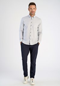 Uomo in piedi con le mani in tasca, che indossa una camicia buttoned grigia chiara, pantaloni scuri e sneaker bianche su uno sfondo neutro.
