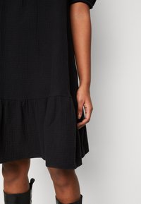 Černé šaty z texturované tkaniny s rozšířeným volánovým lemem. Rukávy jsou nařasené a střih je volný. Ruka je viditelná, jak drží šaty.