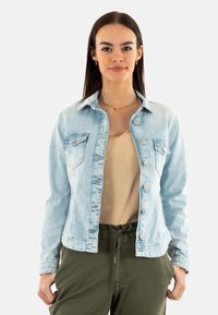 Veste en denim bleu clair avec fermeture à boutons, deux poches poitrine et col classique. Portée par-dessus un haut beige et un pantalon vert. Texture lisse.