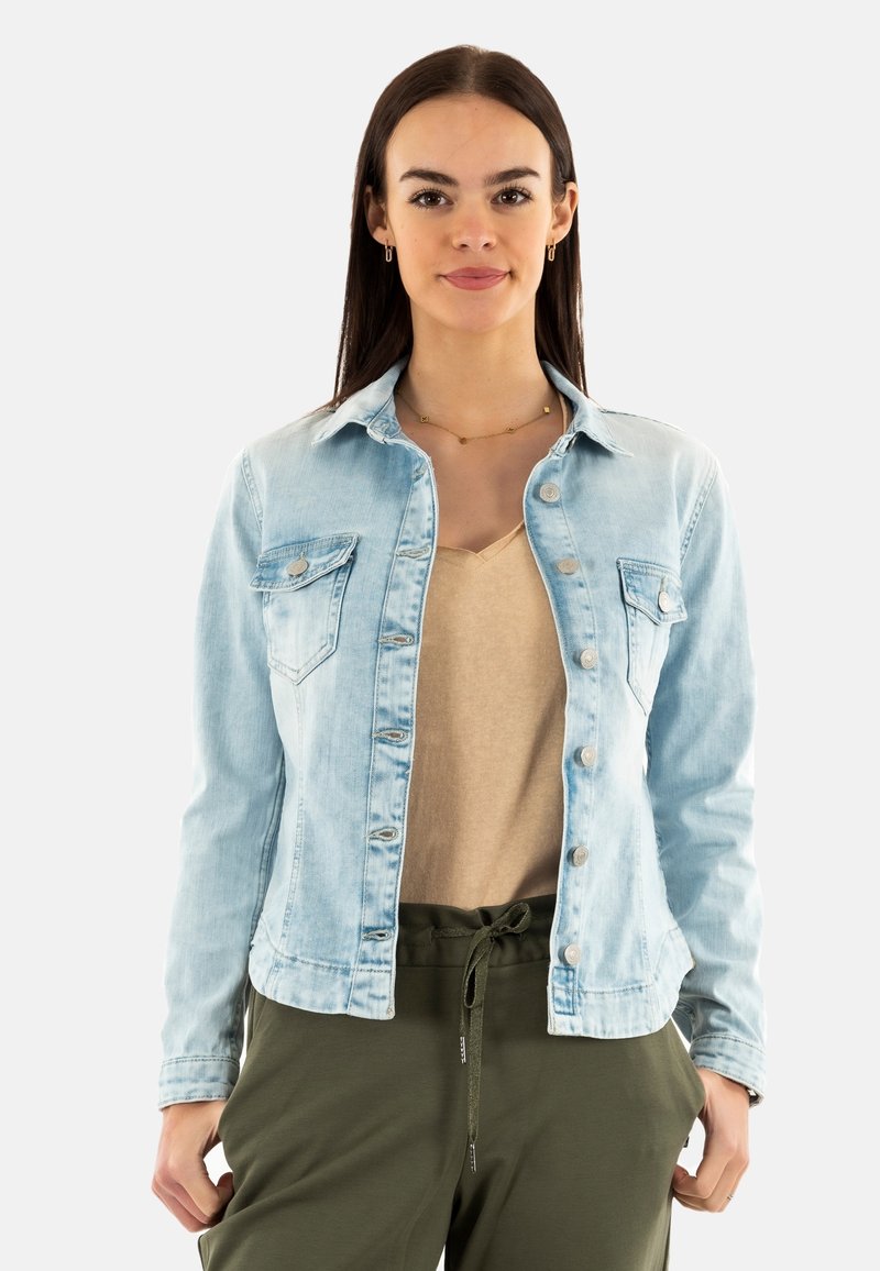 Veste en denim bleu clair avec fermeture à boutons, deux poches poitrine et col classique. Portée par-dessus un haut beige et un pantalon vert. Texture lisse.