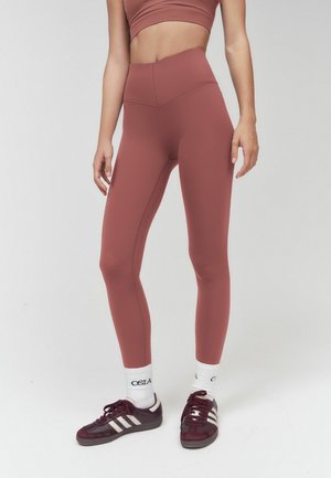 Kobieta nosząca legginsy w kolorze mauve z wysokim stanem, dopasowany sportowy biustonosz, białe skarpetki z czarnym napisem i bordowe sneakersy z białymi paskami.